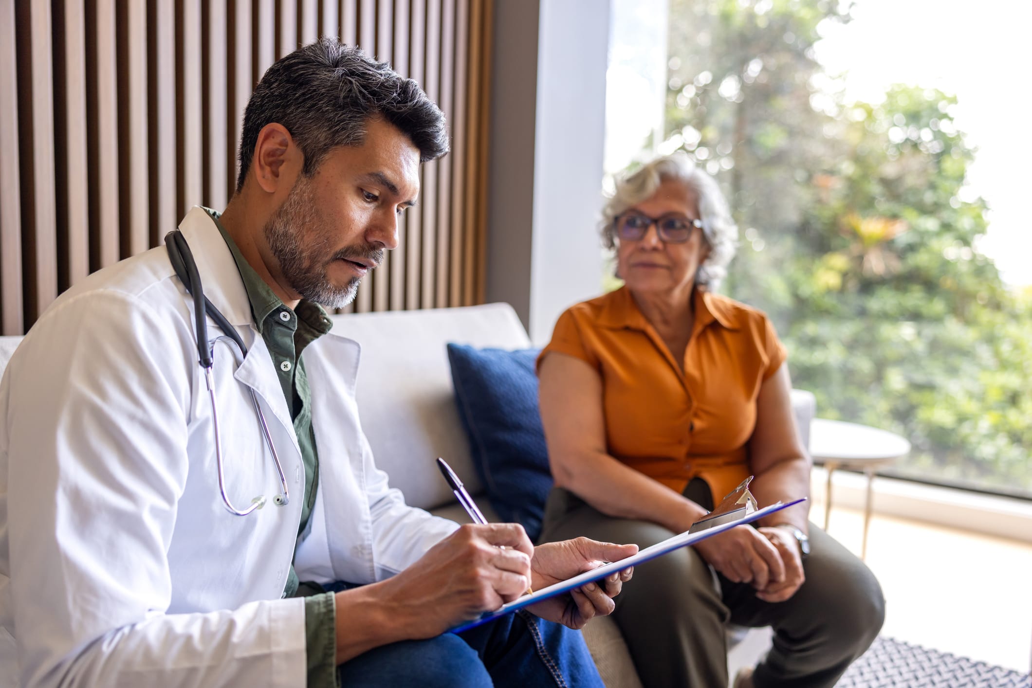 Doctor writing on a clipboard with patient in the background