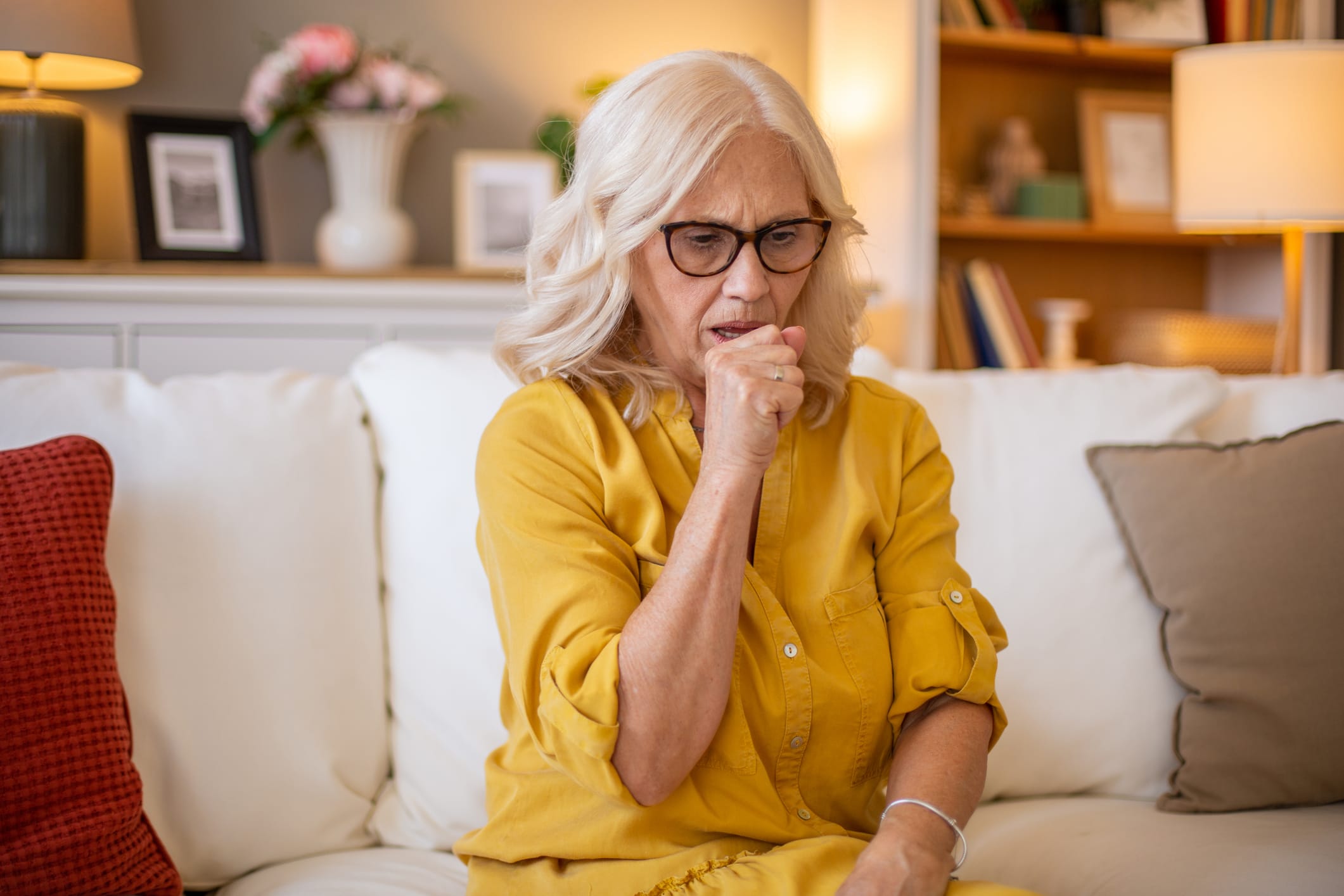 Senior woman suffering from cough, experiencing respiratory problems while sitting on a sofa in her living room