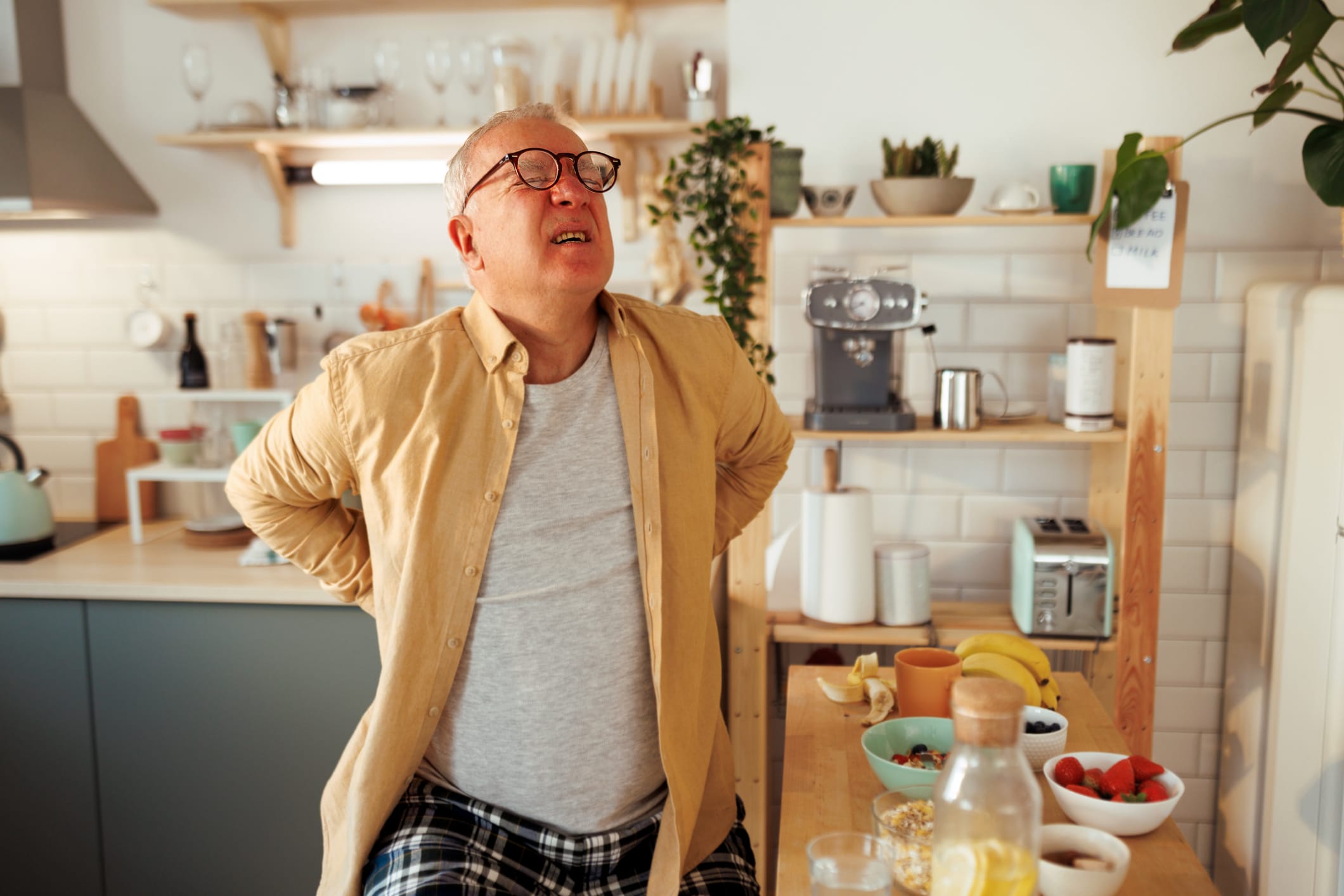 Casually clothed senior man experiencing lower back pain in a domestic kitchen while having a breakfast