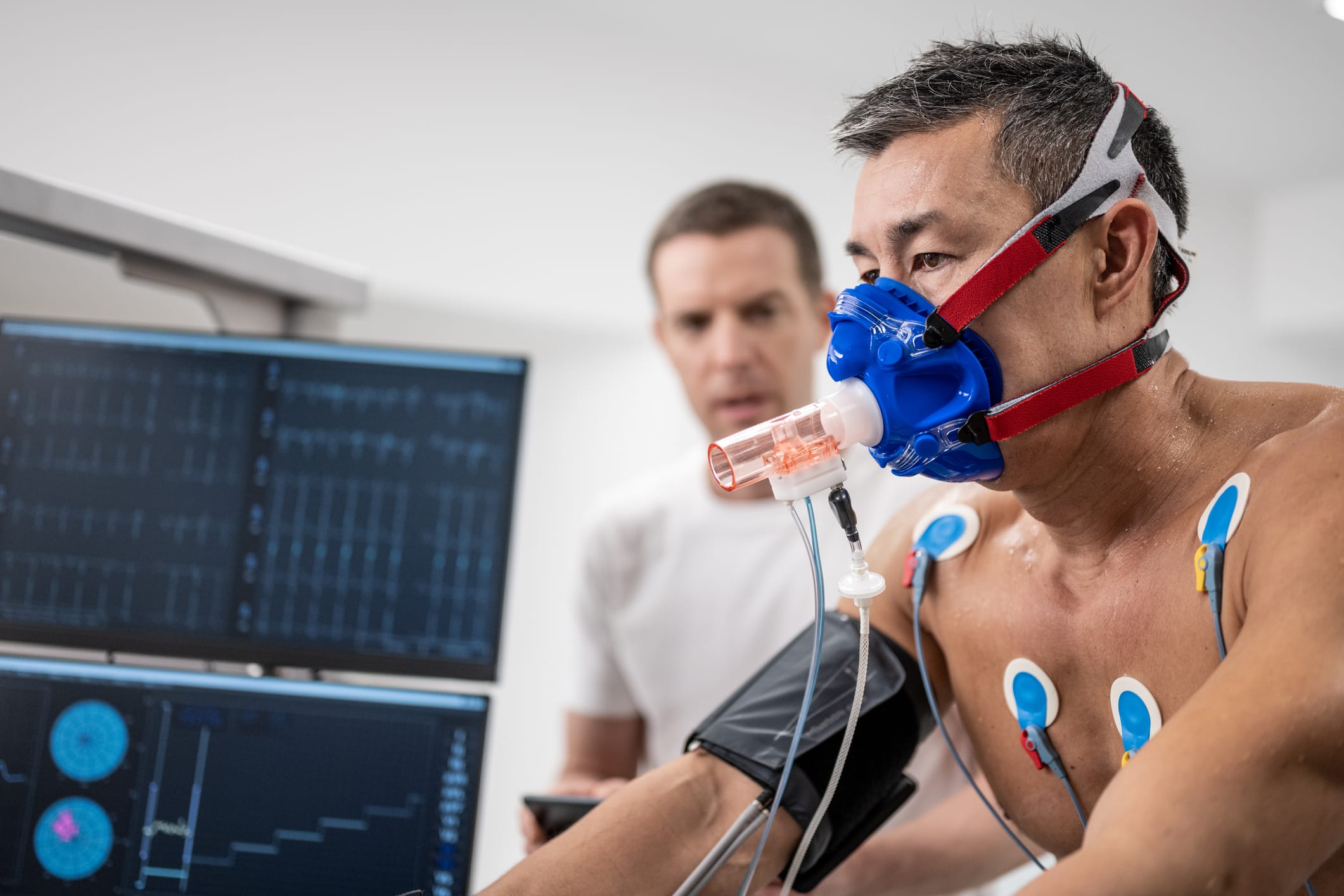 Male doctor observing the progress of a cardiopulmonary stress test taken by the man riding a bicycle ergometer.