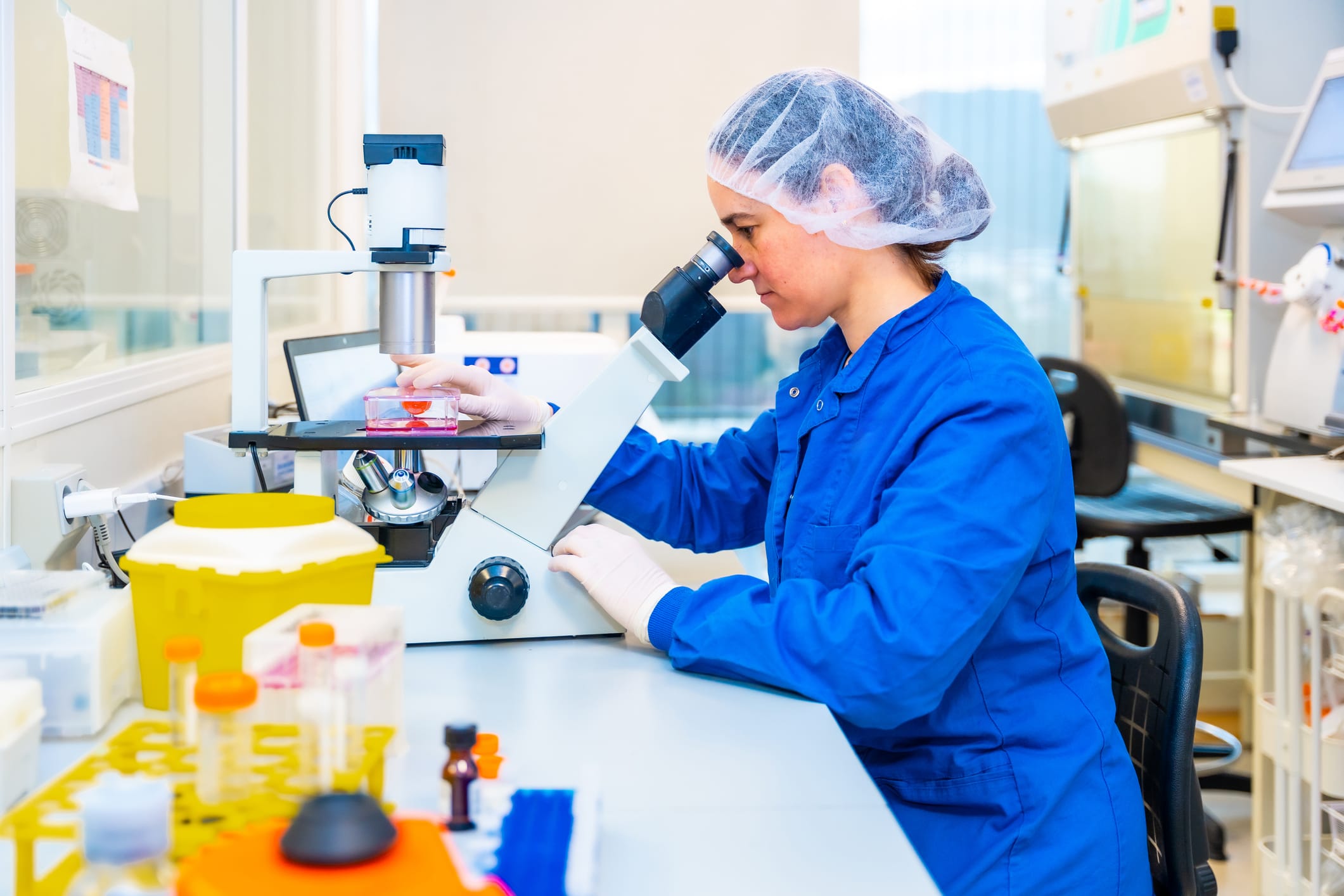 Side view of a female scientist wearing protective gear using a microscope sitting on a research laboratory desk