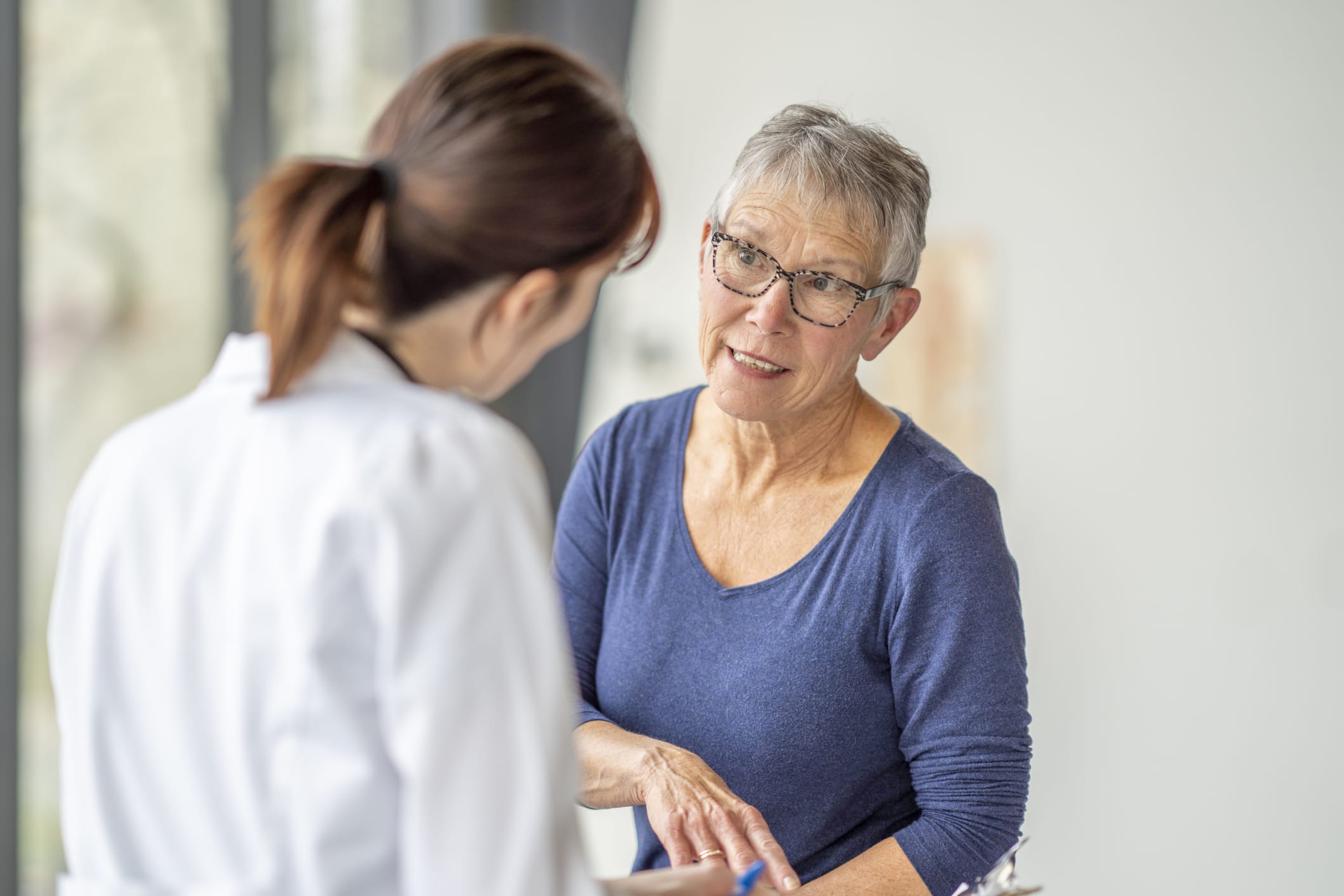 A senior woman who is dressed casually, sits up on an exam table as she attends a health check-up with her doctor. She has a neutral expression on her face as she talks with the female doctor. The doctor is seated across from her and has a clipboard in her hands as she takes notes throughout the appointment.