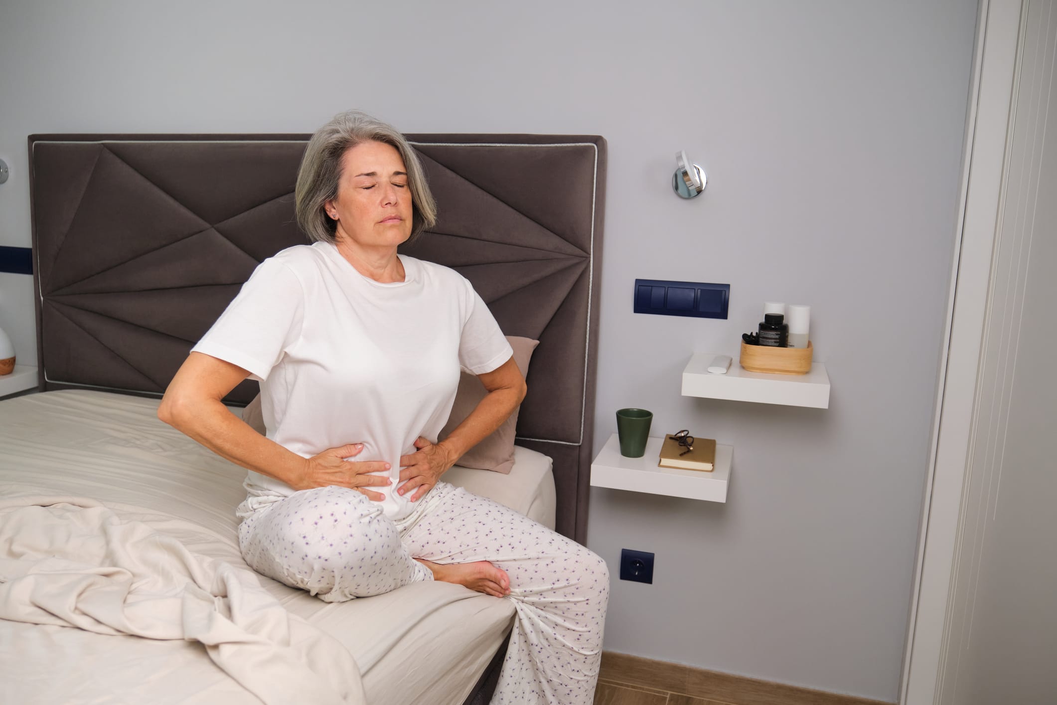 Person sitting on bed holding stomach in pain at home interior